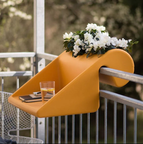 Auf einem leuchtend orangefarbenen Balkontisch stehen ein Glas Wasser, ein Notizbuch und ein Topf mit weißen Blumen und verschönern den Außenbereich.