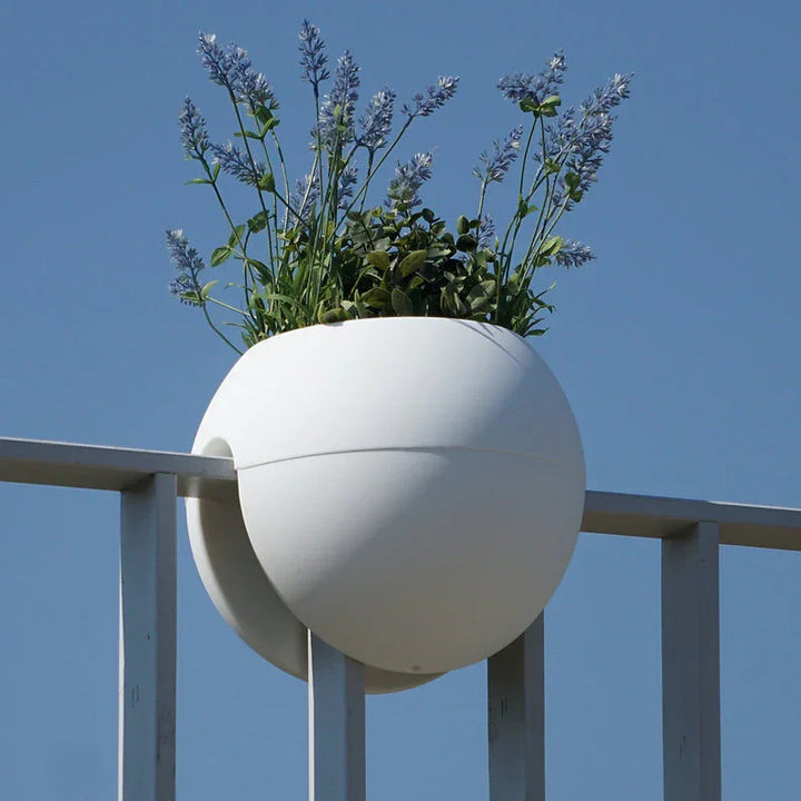 Ein runder weißer Blumentopf mit violetten Blüten ist an der oberen Stange eines Metallzauns vor einem klaren blauen Himmel befestigt